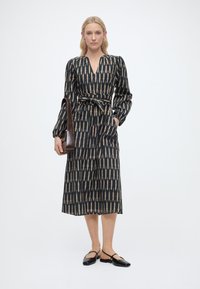 Γυναίκα με midi φόρεμα μακρυμάνικο με σχέδια και ζώνη στη μέση, μαύρα επίπεδα παπούτσια και καφέ τσάντα, στέκεται μπροστά σε απλό λευκό φόντο.