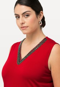 Červený top bez rukávů s výstřihem do V, zdobený jemným černým a stříbrným korálkovým lemem podél výstřihu. Hladká, jemná textura látky.