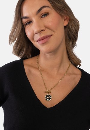 Frau mit hellbraunem Haar, die ein schwarzes V-Ausschnitt-Oberteil und eine goldene Kettenhalskette mit einem schwarzen, kreisförmigen Anhänger trägt, der ein zentrales Auge und Halbmonde zeigt.