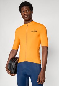 Maillot de cyclisme orange à manches courtes avec une fermeture éclair sur le devant et un logo noir. Un homme tient un casque noir, portant un short de cyclisme bleu marine.