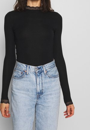 Kobieta ubrana w czarną bluzkę z długim rękawem z koronkowym wykończeniem przy dekolcie i mankietach, zestawioną z jasnoniebieskimi, wysokim stanem jeansami.