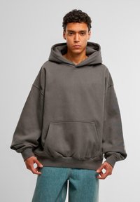 Hoodie oversize grigio scuro in morbido tessuto con tasca frontale e polsini a costine. Presenta un logo sottile sul petto e strisce in tonalità tenui sulle maniche.
