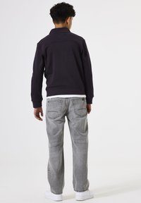 Joven de pie de espaldas, vistiendo una chaqueta oscura, jeans gris claro, zapatillas blancas y una camiseta blanca debajo de la chaqueta.