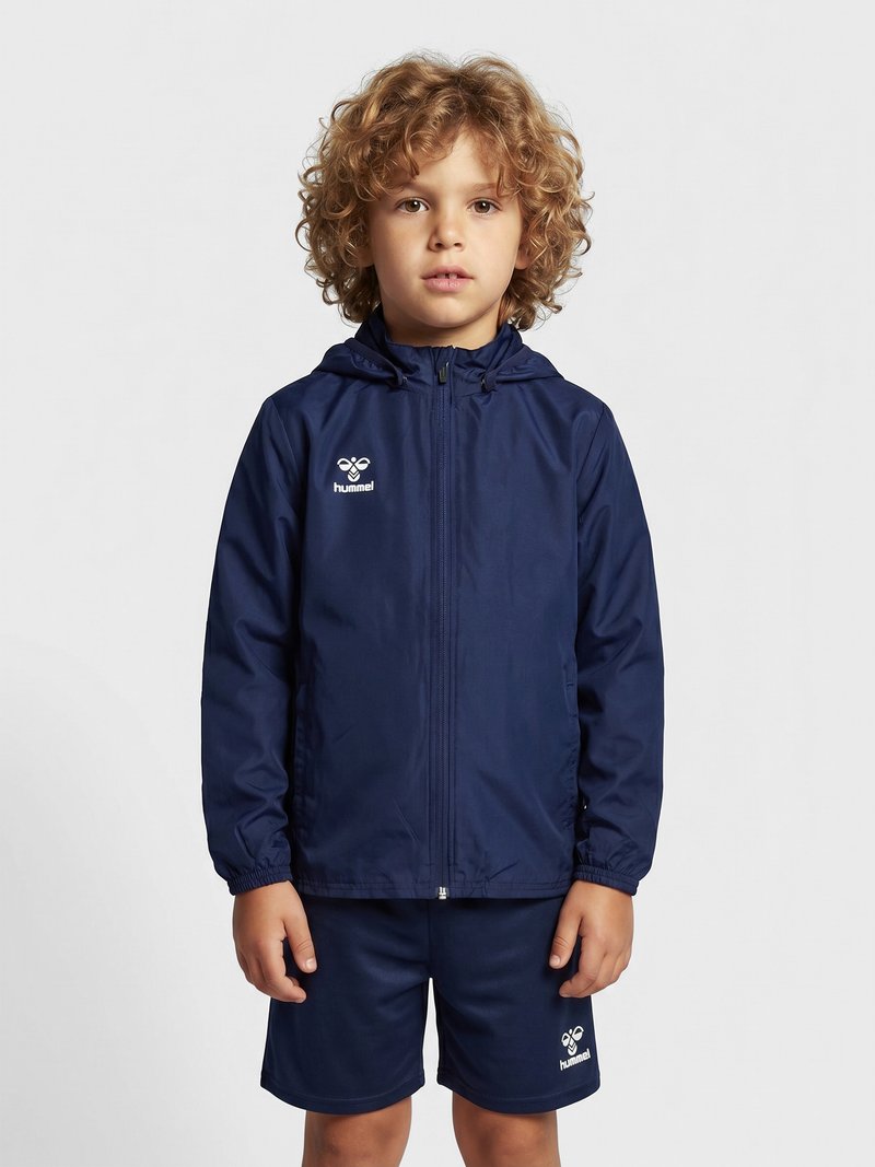 Kind met krullend haar, gekleed in een marineblauwe jas met rits en bijpassende korte broek, staand tegen een effen lichte achtergrond.
