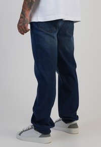 Jeans de mezclilla azul oscuro de corte holgado con bolsillos traseros, usados con zapatillas de deporte de patrón gris y blanco. Tejido texturizado visible.