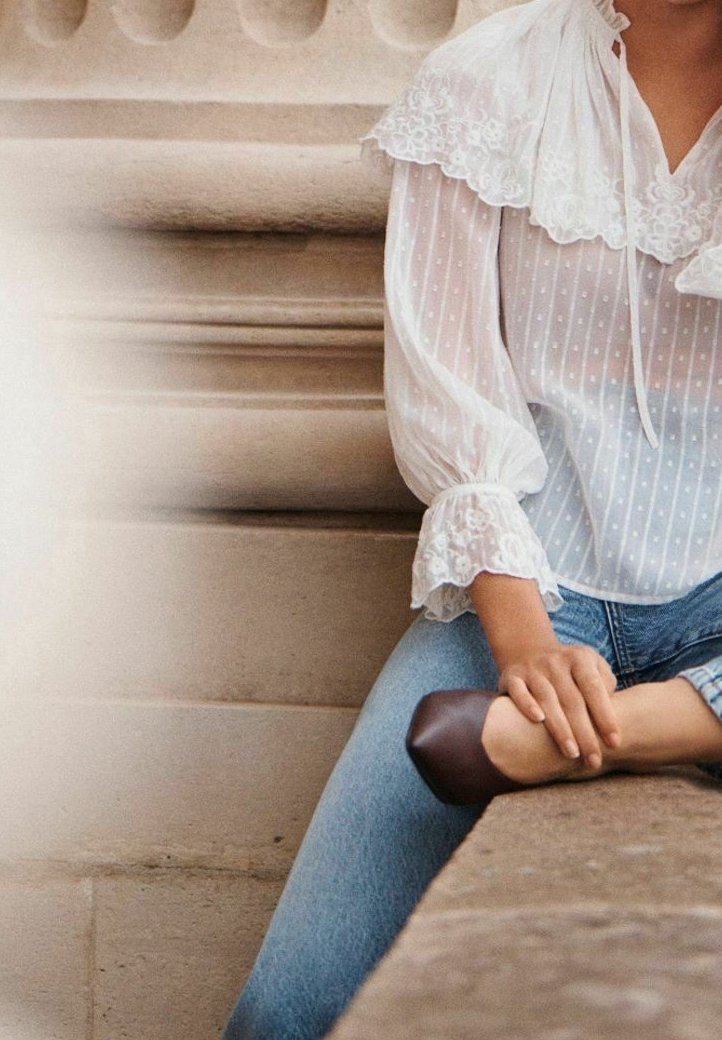 Personne portant un chemisier blanc transparent avec des détails en dentelle et un jean bleu, assise sur des marches en pierre, tenant un pied avec une chaussure plate marron.