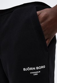 Χέρι στην τσέπη μαύρου παντελονιού με λευκό λογότυπο Björn Borg Stockholm 1956 στο μηρό.