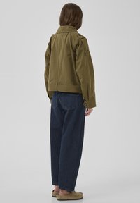 Person trägt eine olivgrüne, verkürzte Jacke, dunkelblaue Jeans mit weitem Bein und beige Slip-On-Schuhe, steht mit dem Rücken zur Kamera vor einem schlichten grauen Hintergrund.