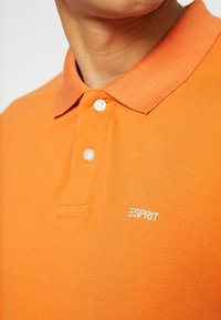 Polo arancione con tessuto testurizzato, patta con tre bottoni e logo "ESPRIT" ricamato in bianco sul petto.