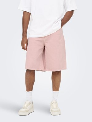 Homme portant un short rose clair jusqu’aux genoux, des chaussettes blanches, des baskets crème et un t-shirt blanc ample, debout avec une main dans la poche.