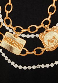 Svart tyg med gyllene kedjedesigner och vita och silverpärlor, inklusive en guldtagg märkt "MOSCHINO" och en björntroll.