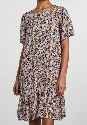 Frau trägt ein knielanges, geblümtes Kleid mit kurzen Ärmeln und einem gerafften Saum in Pink, Braun und Schwarz auf weißem Hintergrund.