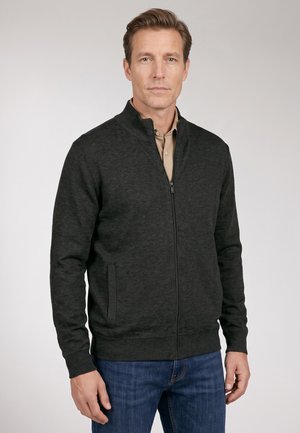 Mann trägt einen dunkelgrauen Reißverschluss-Pullover über einem beigen Hemd und blauen Jeans, steht vor einfachem hellem Hintergrund.