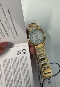 Citizen-horloge in goudkleur met een zilverwitte wijzerplaat, uurmarkeringen en datumweergave. Metalen band met rechthoekige schakels. Instructiekaart zichtbaar.