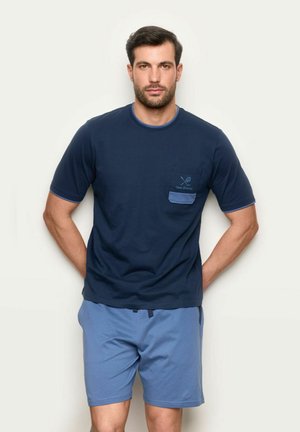 Mężczyzna stojący z rękami za plecami, ubrany w granatowy T-shirt z kieszonką i jasnoniebieskie szorty na jednolitym tle.