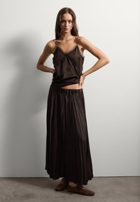 Bruine satijnen camisole met kantdetails, gecombineerd met een geplooide bruine rok. Zachte textuur, vloeiende vorm en een elastische tailleband. Bruine schoenen.