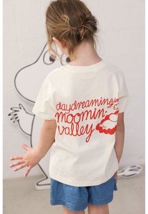 Fonott hajú gyerek fehér pólóban, amelyen piros betűkkel az áll: „álmodozás a Mumin-völgyben”, kék rövidnadrágban, egy falfestmény mellett áll.