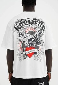 White t-shirt featuring a large graphic on the back with stylized text, an eagle, and a heart, accented by black and red details.