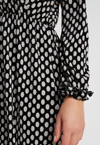 Schwarzes und weißes gepunktetes Kleid aus strukturiertem, plissiertem Stoff. Verfügt über einen gerafften Taillenbund und Rüschensaum an den Ärmeln.