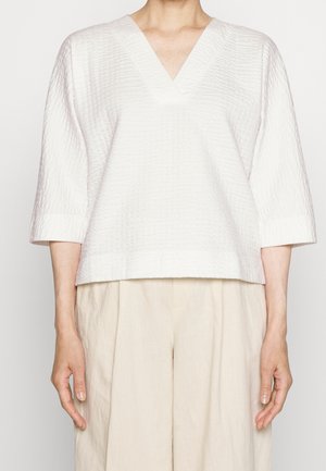 Femme portant un haut blanc texturé à col en V avec des manches trois-quarts et un pantalon beige ample, debout les bras détendus le long du corps.