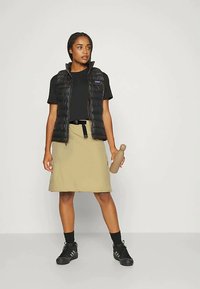 Colete acolchoado preto sobre uma t-shirt preta, combinado com uma saia bege. Sapatos pretos e uma garrafa de água castanha estão segurados em uma mão.