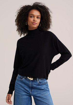 Frau mit lockigem Haar, die goldene Creolen, einen schwarzen langärmeligen Rollkragenpullover und blaue Jeans mit Gürtel trägt.