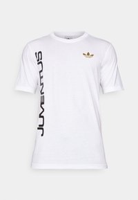 Camiseta de algodón blanca con mangas cortas. Presenta texto vertical en negro "JUVENTUS" y un logo dorado de Adidas en el pecho. Diseño simple.
