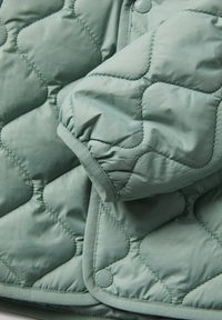 Quiltet jakke i dæmpet grøn, med en blød tekstur, samlede manchetter og kontrasterende kantbinding. Bemærkelsesværdig rund logo detalje til stede.