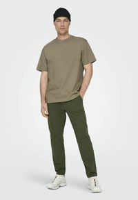 Camiseta caqui y pantalones verde oliva. El modelo lleva un gorro negro y zapatillas blancas. Diseño sencillo, mangas cortas, tela ligera.