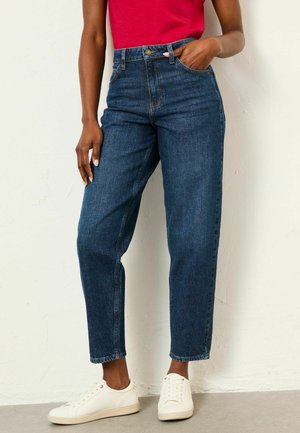 Person iført højtaljede mørkeblå jeans, rød top og hvide sneakers, stående med den ene hånd i lommen mod en ensfarvet væg.