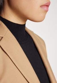 Gros plan d'une personne portant un pull col roulé côtelé noir et un blazer beige, montrant la partie inférieure du visage et le cou.