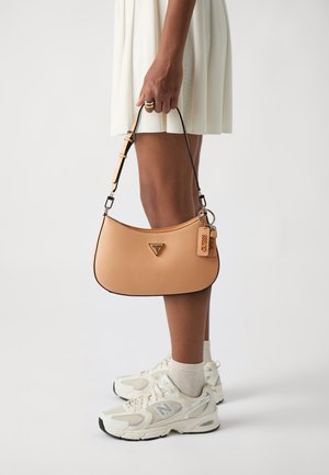Tan designer handbag with a curved shape, textured surface, black trim, and a keychain. Paired with white sneakers and a white pleated dress.