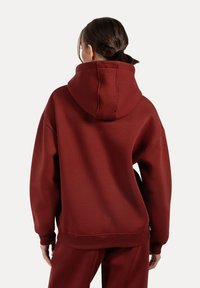 Felpa oversize bordeaux con cappuccio grande, polsini e orlo a coste, realizzata in tessuto morbido, che mostra una superficie liscia sul retro.