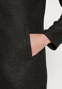 Mörkgrå ullkappa med sidofickor och avslappnad ärmdesign, som visar en mjuk textur och subtil struktur.