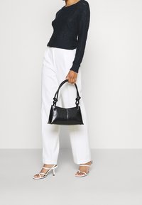 Černá kožená kabelka s strukturovaným tvarem, dvěma uzlovými rukojeťmi a hladkou texturou, spárovaná s přiléhavým tmavě modrým svetrem a bílými kalhotami.