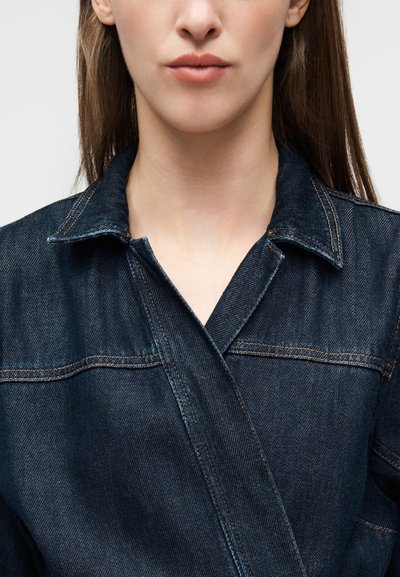 Veste en denim foncé avec col et design croisé, détails de coutures visibles et texture de poids moyen.
