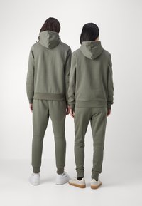 Zwei Personen tragen olivgrüne Kapuzenpullover und passende Jogginghosen, die über gerippte Bündchen und einen weichen, strukturierten Stoff verfügen.