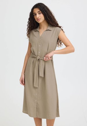 Kobieta z długimi kręconymi włosami, ubrana w beżową sukienkę bez rękawów zapinaną na guziki, z związanym paskiem, stojąca na jednolitym jasnym tle.