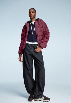 Bordeauxkleurige pufferjack met een capuchon, gelaagd over een marineblauwe hoodie met rits en een witte top. Losse donkere denimbroek en bruine sneakers maken de look compleet.