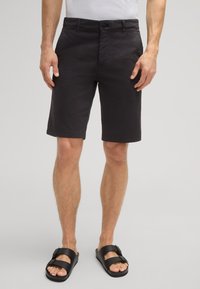 Pantaloni corti grigi scuri in cotone con una texture liscia, dotati di chiusura frontale a bottone e tasche laterali, abbinati a sandali neri con cinturino.