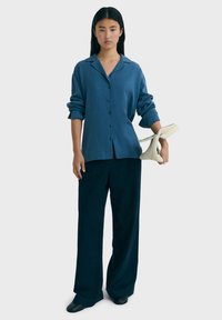 Camisa azul de botones con cuello, mangas enrolladas, combinada con pantalones oscuros de pierna ancha. El modelo sostiene un bolso color crema.