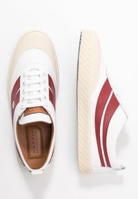 Baskets blanches avec deux bandes rouges latérales, semelle beige texturée, lacets à l'avant et semelle intérieure marron portant la marque "Bally", vues de dessus et de côté.