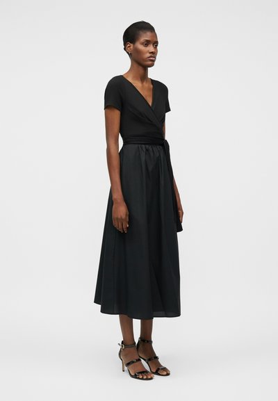 Γυναίκα που στέκεται, φορώντας μαύρο φόρεμα τύπου wrap με κοντά μανίκια και μίντι φούστα, σε συνδυασμό με μαύρα πέδιλα με λεπτά λουράκια και ψηλό τακούνι.