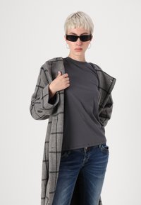 Casaco de lã cinza xadrez com grandes lapelas, sobre uma t-shirt cinza escura, combinado com jeans azuis. Os acessórios incluem óculos de sol pretos e brincos de argola.
