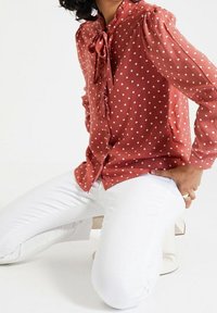 Rustfärgad blus med prickar, rynkad fram, knytkrage och puffärmar, kombinerad med figurnära vita byxor. Slät tygstruktur.