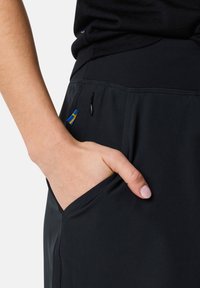 Mustat urheilushortsit, joissa on sivutasku, jossa on vetoketju. Taskussa on sininen ja keltainen korostus, mikä korostaa toiminnallisia suunnitteluelementtejä.