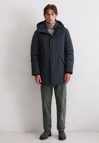 Navyblaue Puffy-Jacke mit Kapuze, ausgestattet mit einem Reißverschluss, seitlichen Taschen und einem strukturierten Design. Kombiniert mit grünen Cordhosen und schwarzen Stiefeln.