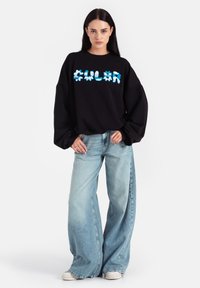 Czarny, oversize sweatshirt z niebieskim napisem graficznym, luźny krój, szerokie nogawki w jasnoniebieskim denimu, w połączeniu z beżowymi trampkami.