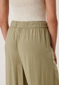 Szerokie spodnie w kolorze jasnej oliwkowej zieleni, z elastycznym, marszczonym paskiem w talii i miękką teksturą tkaniny. Prosty i minimalistyczny design bez widocznych wzorów.