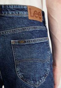 Tummansiniset farkut, joissa on teksturoitu denim-viimeistely. Sisältää ruskean nahkapatchin, jossa on "Lee"-logo, sekä pienen logo-tunnisteen takataskussa.